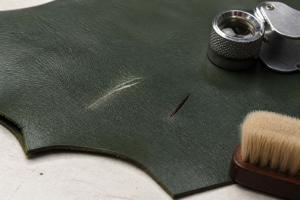 Dark green leather test panel with a shallow finish scuff and a short deeper cut beside a loupe